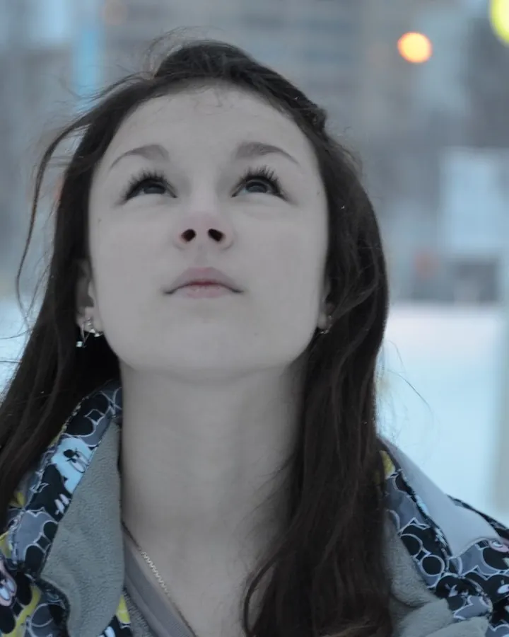 Femme en veste à motif, debout dans la neige, regardant vers le haut, avec des lumières floues en arrière-plan.
