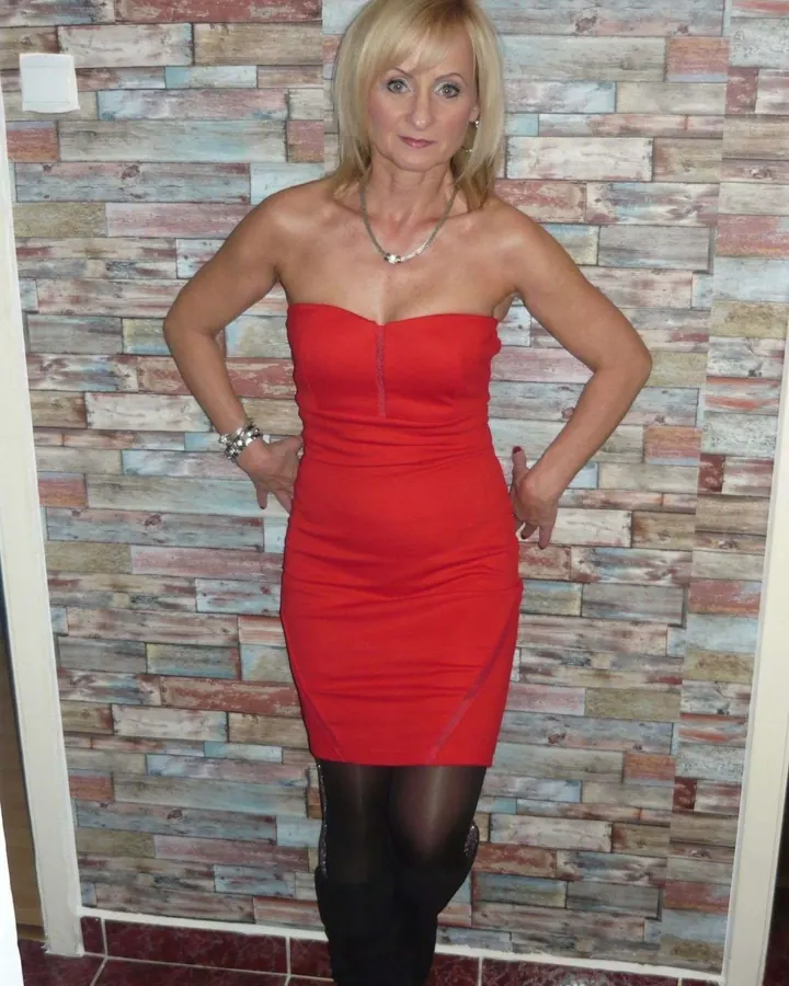 Femme blonde en robe rouge ajustée, bas noirs et bottes, pose les mains sur les hanches devant un mur en bois.