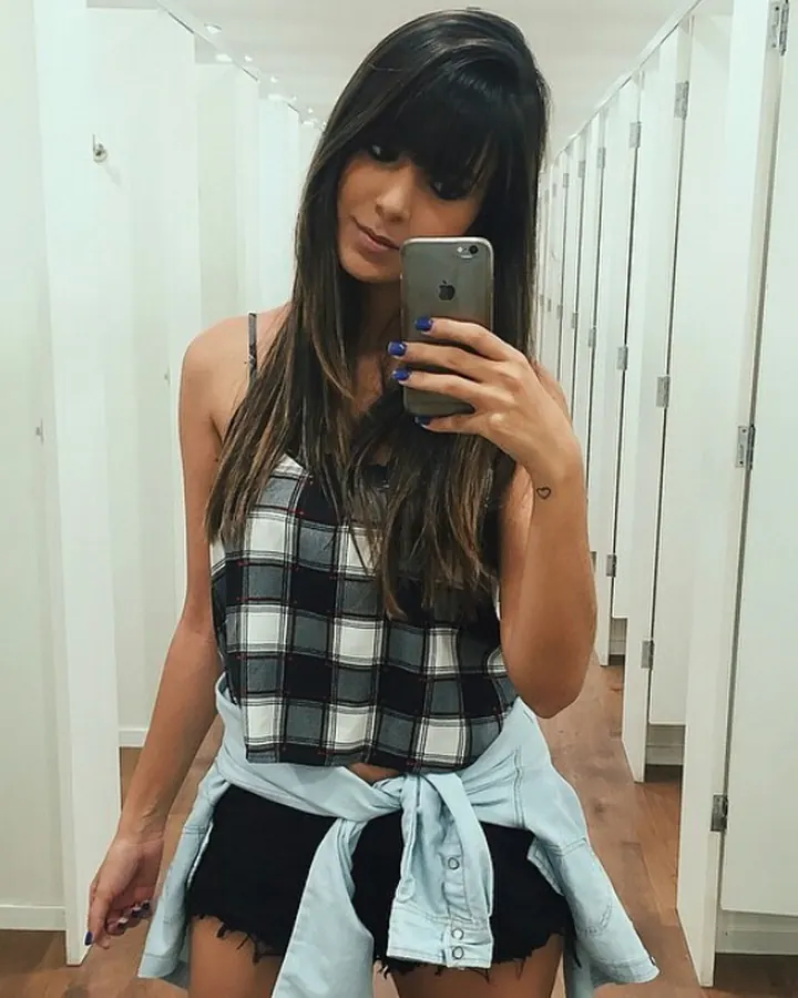 Jeune femme aux cheveux bruns foncés en haut à carreaux, posant dans un vestiaire, tenant un téléphone.