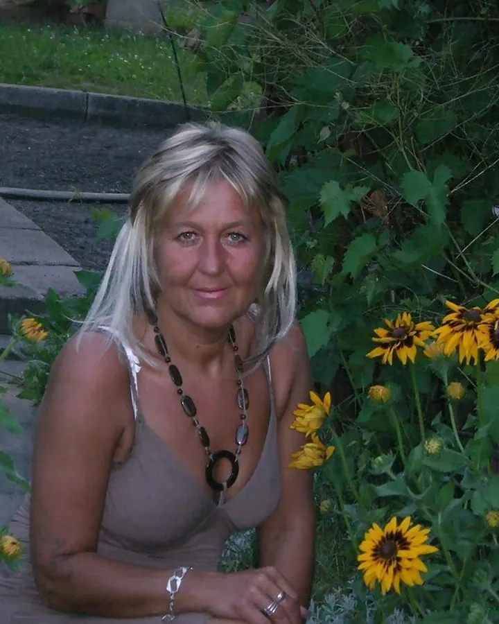Femme blonde assise parmi des fleurs jaunes, portant un haut beige et un collier