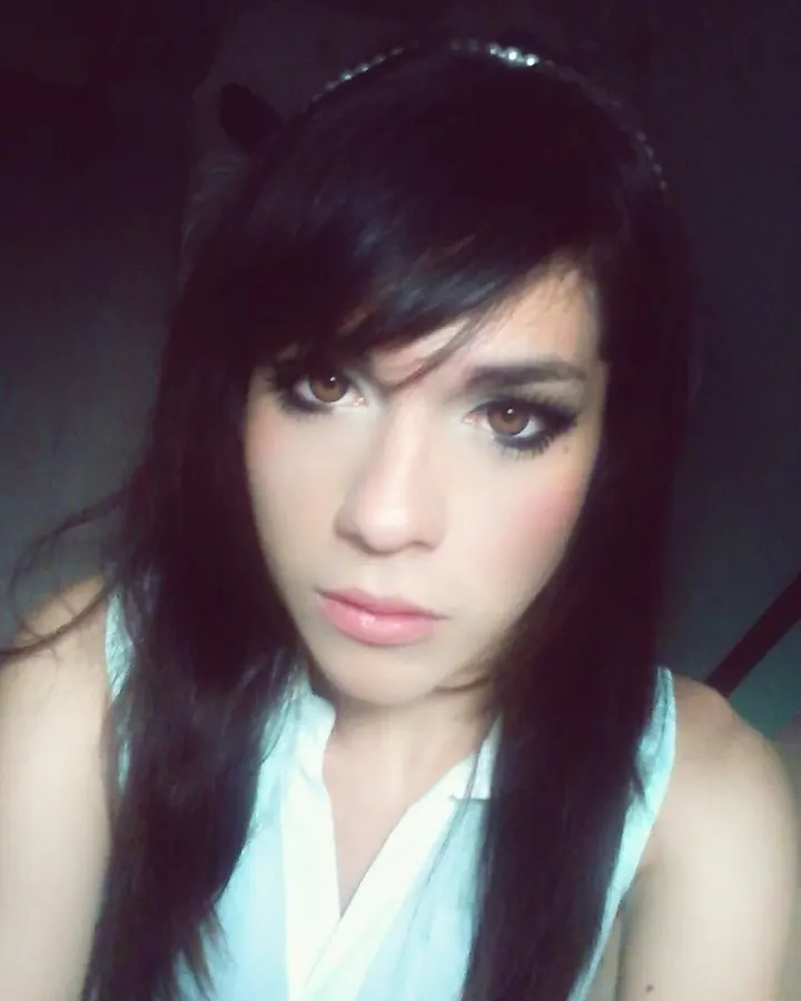 Portrait en gros plan d'une femme aux cheveux noirs longs et maquillage prononcé, portant un haut blanc sans manches.
