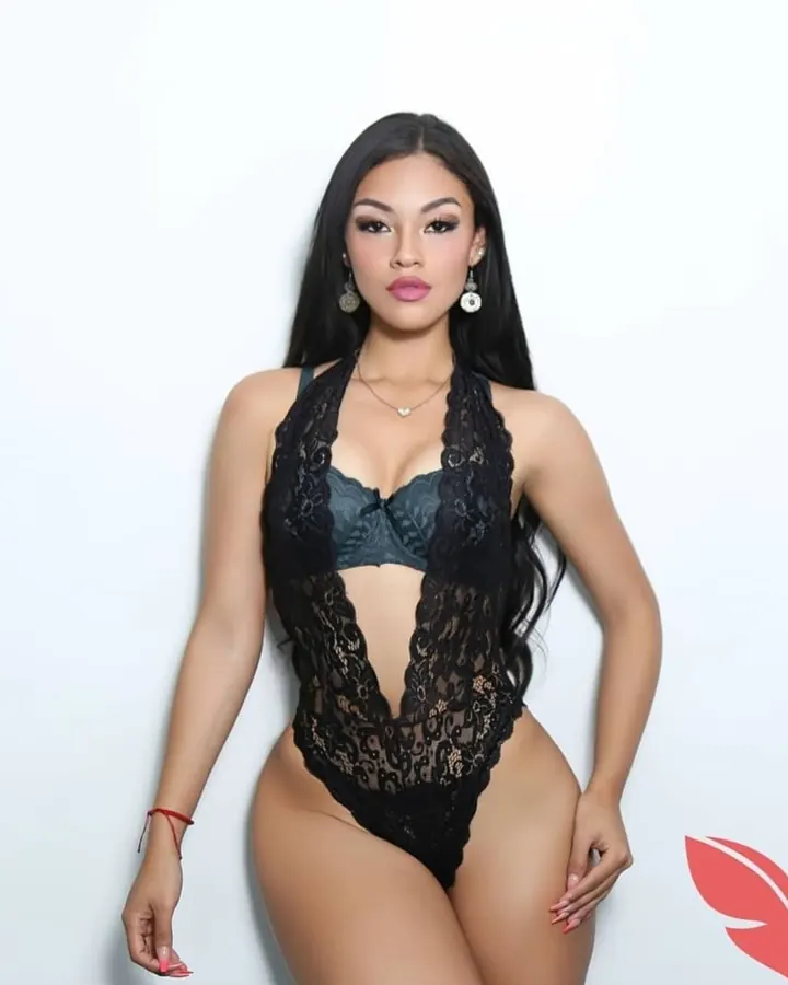 Femme en lingerie noire à dentelle, portant un collier, regardant directement l'appareil, devant un fond blanc.