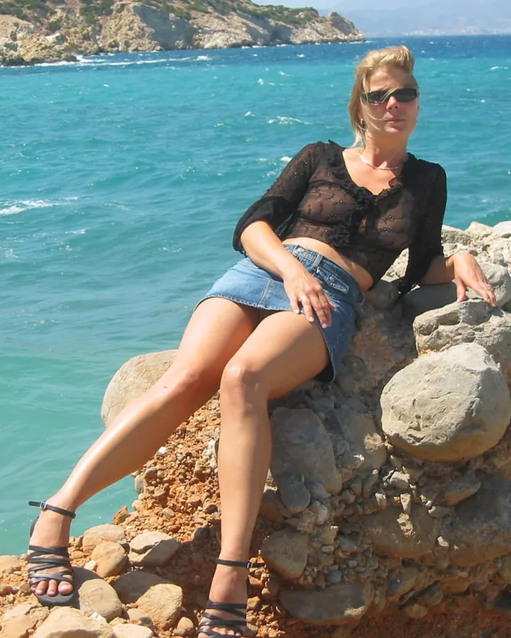 Femme blonde aux yeux marrons, assise sur un mur de pierres près de la mer, portant une chemise noire transparente et un short en jean.