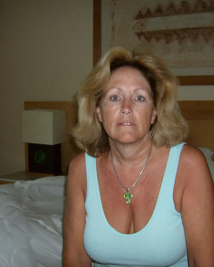 Femme aux cheveux blonds, assise sur un lit, portant un haut bleu clair, regardant directement l'appareil.