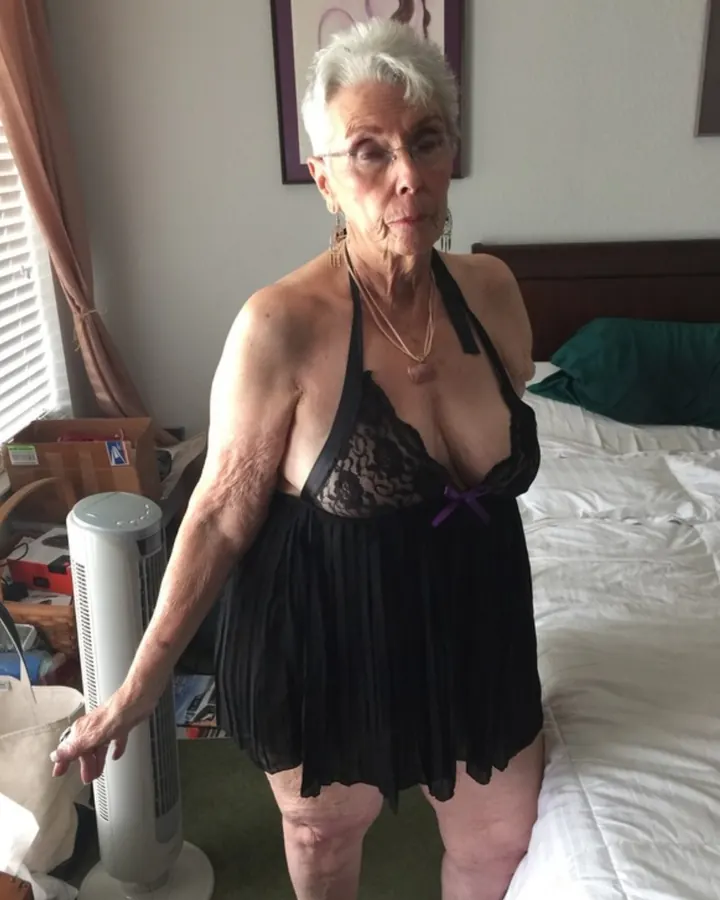 Femme aux cheveux blancs, portant un haut noir et un short noir, debout dans une chambre.