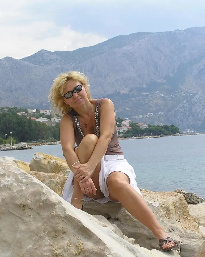 Femme blonde aux cheveux courts, assise sur des rochers près d'une mer, portant un haut marron, jupe blanche, lunettes de soleil, regardant vers l'appareil.