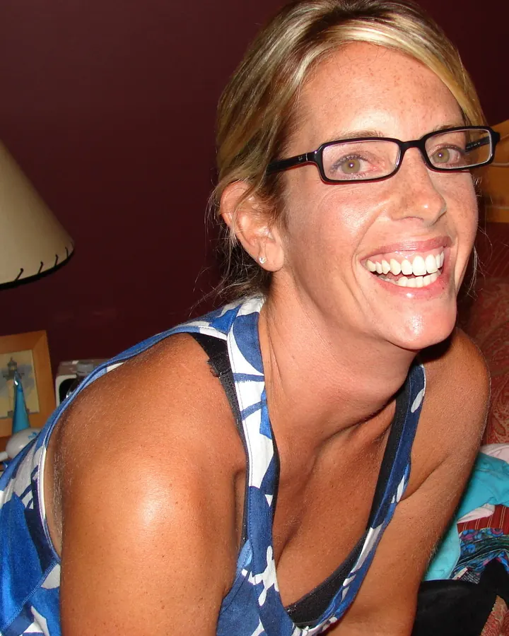 Femme aux cheveux blonds, lunettes, souriant largement, vêtue d'un haut bleu et blanc, dans un intérieur chaleureux.