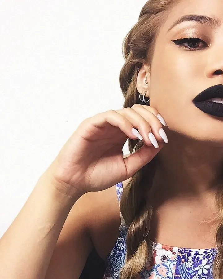 Femme aux cheveux blonds et yeux marrons en gros plan, maquillage prononcé, lèvres noires, main près du visage.