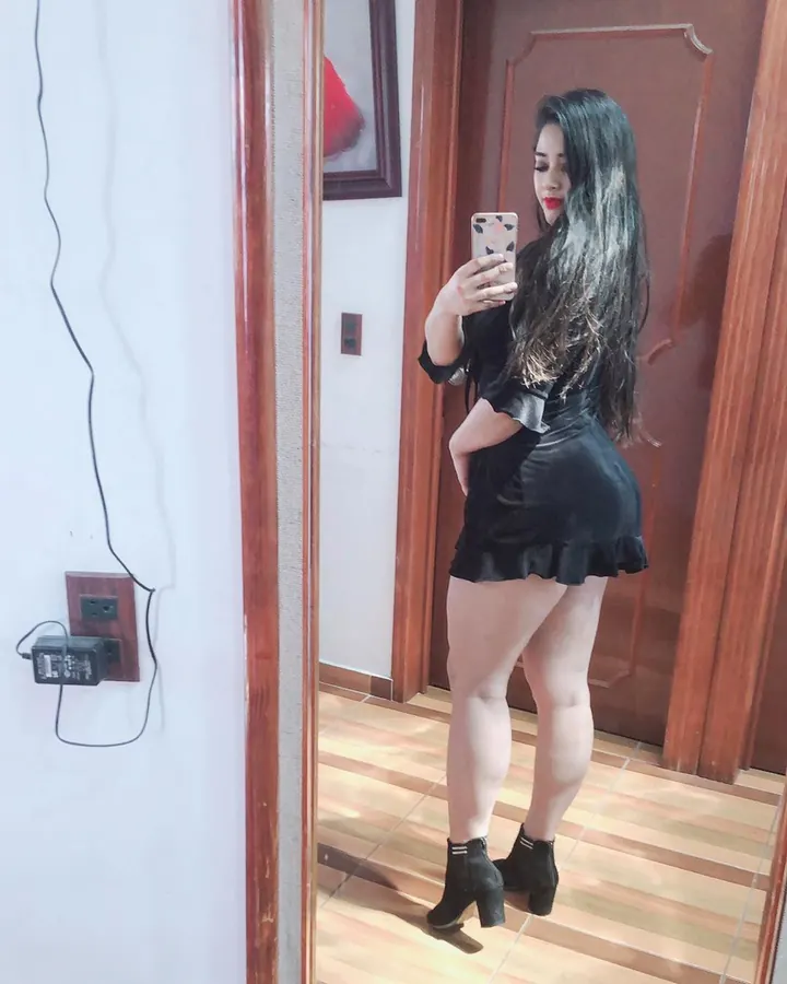 Femme aux cheveux longs bruns foncés, portant une robe noire courte avec volants, prenant un selfie devant un miroir, pieds chaussés de bottines noires.
