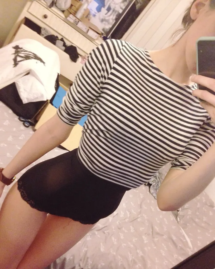 Jeune femme aux cheveux châtains portant un haut rayé noir et blanc et une jupe noire, prenant un selfie devant un miroir dans une chambre.