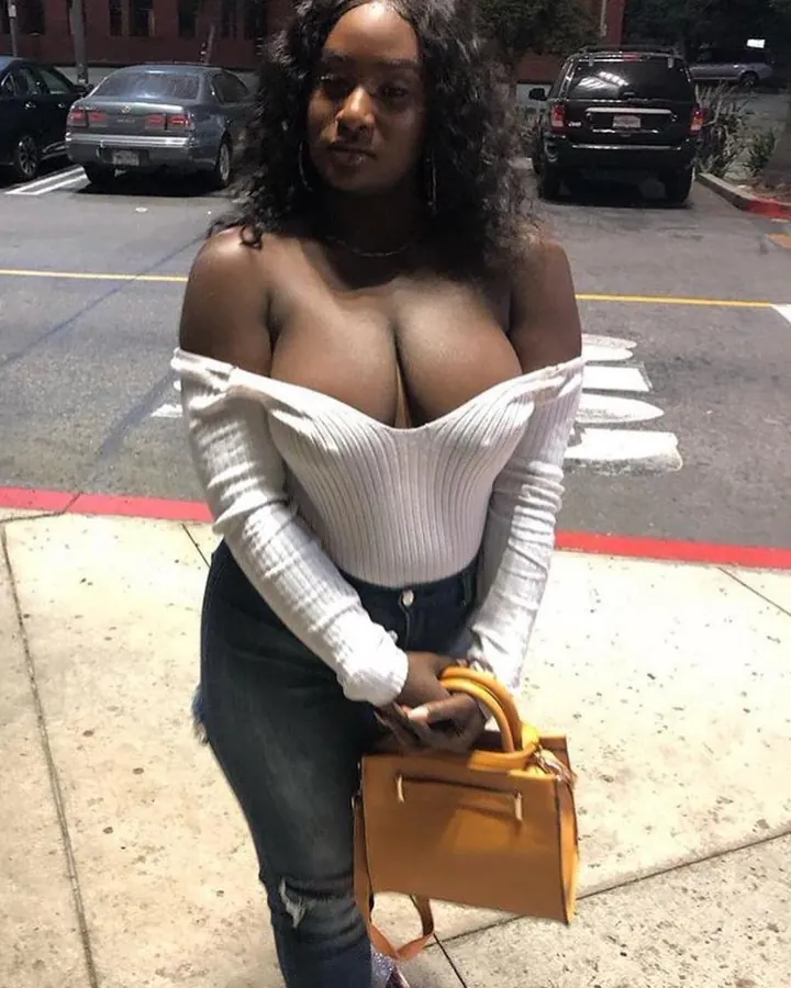Femme aux cheveux bruns foncés, portant un haut blanc à manches longues, tenant un sac jaune, debout sur un trottoir, regardant l'appareil.