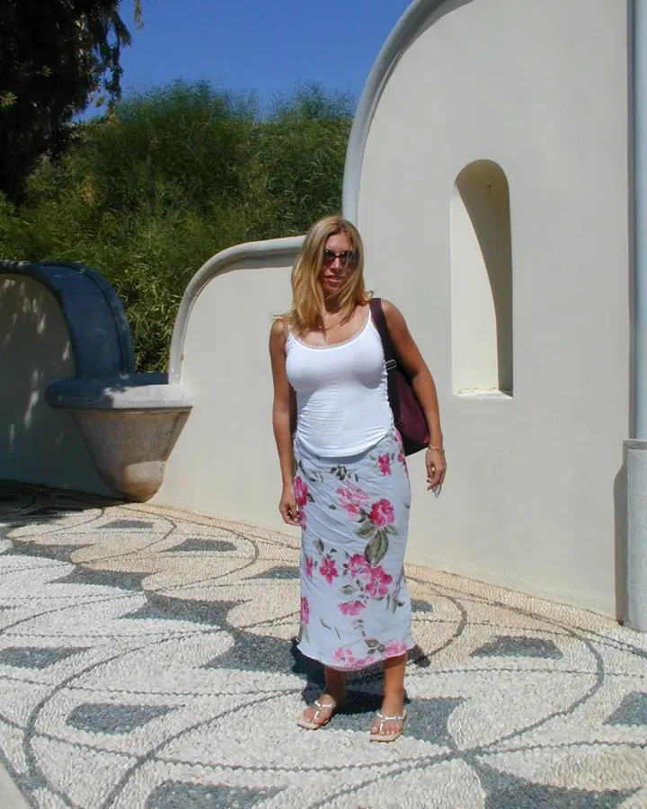 Femme aux cheveux blonds platine, portant un haut en latex noir et un pantalon assorti, tenant les mains sur les hanches, regardant directement l'appareil.