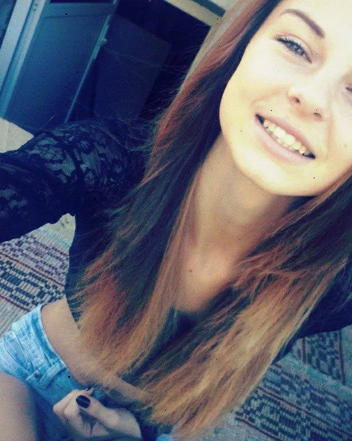 Jeune femme aux cheveux châtains, souriante, en haut noir ajouré et shorts bleus, prenant un selfie dans une pièce intérieure.