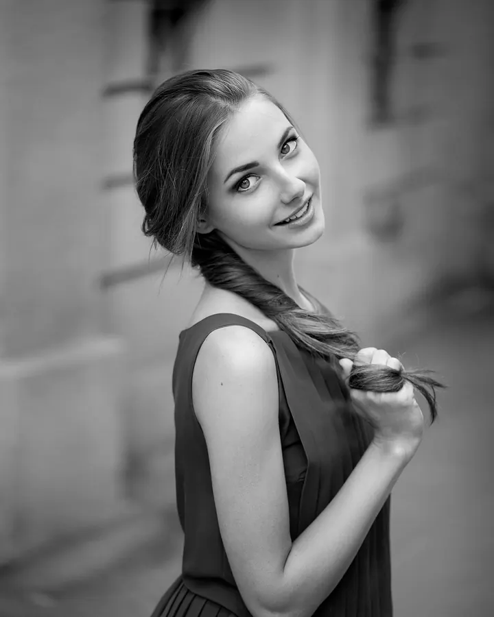 Jeune femme blonde en robe noire, vue de profil, tenant sa longue tresse, souriant doucement devant un fond flou de bâtiment ancien.