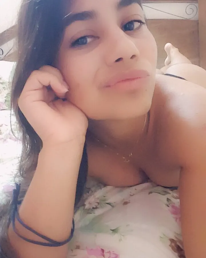 Femme aux cheveux bruns foncés, souriante, en haut floral, posant allongée sur un lit avec un bras derrière la tête.