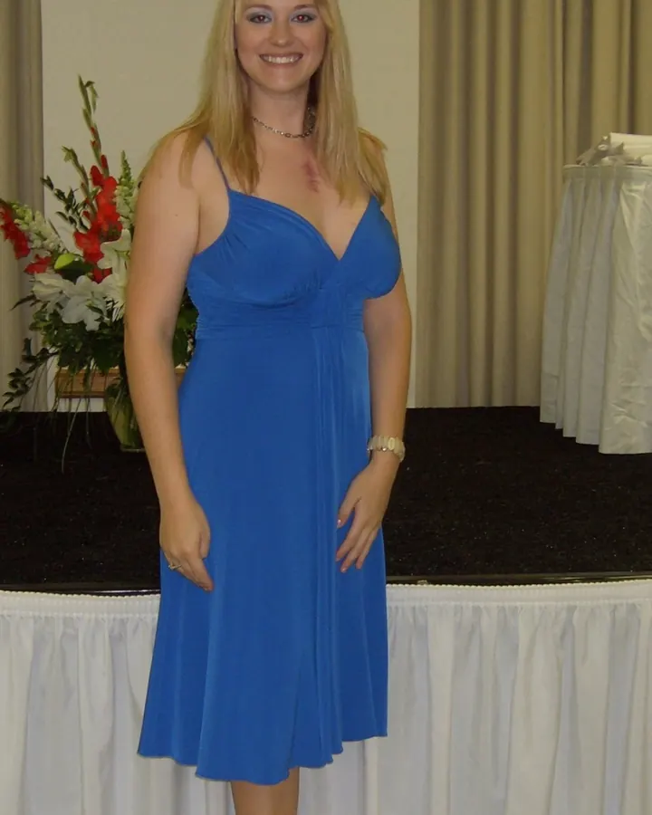Femme blonde en robe bleue à bretelles, souriant, debout près d'une table avec des fleurs.