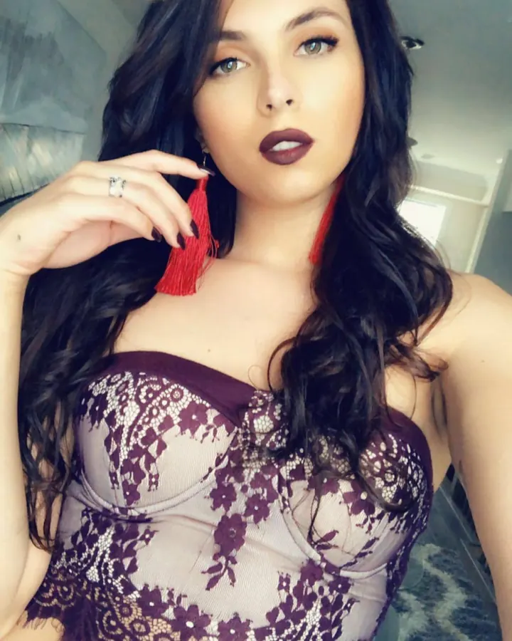 Femme brune en haut violet à dentelle, tenant un bijou rouge, regardant l'appareil photo.