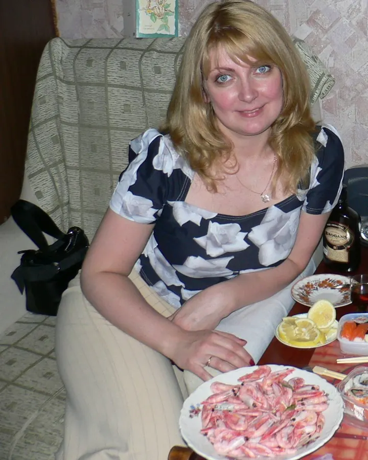 Femme blonde souriante, assise à table avec des crevettes et du sushi, portant une robe à fleurs blanches et bleues.