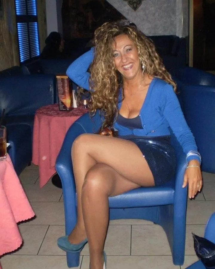 Femme aux cheveux blonds, en robe noire, souriant à l'appareil photo dans une pièce lumineuse.
