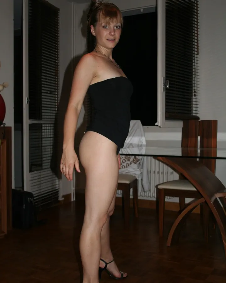 Femme blonde aux cheveux courts, portant un haut noir sans bretelles et des sandales noires à talons, debout dans une pièce avec un sol en parquet et une table en verre.