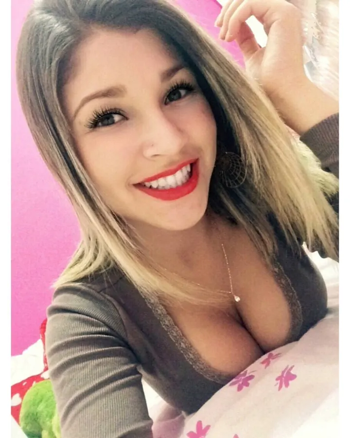 Femme blonde aux cheveux longs, portant un haut noir, souriant à la caméra, avec un piercing au nez et des yeux sombres.