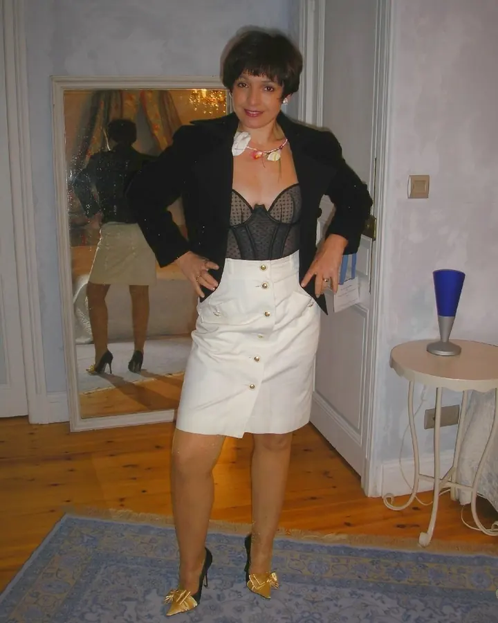 Femme aux cheveux bruns noirs en corset noir et jupe blanche boutonnée posant devant un miroir, mains sur les hanches