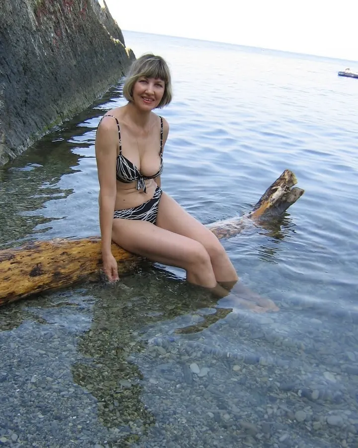 Femme blonde en maillot de bain noir et blanc assise sur un tronc dans l'eau, souriante