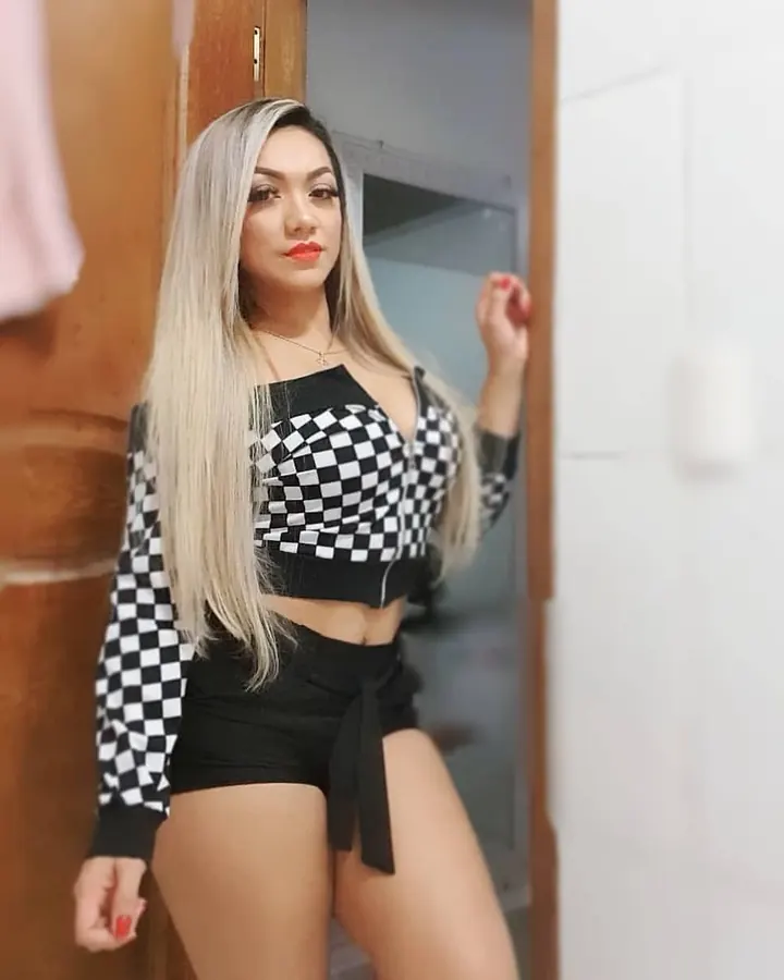 Femme aux cheveux longs blonds platine, top damier noir et blanc, short noir noué, main levée contre une porte