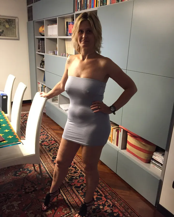 Femme aux cheveux blonds, en pull bleu, posant de face contre un mur en briques.