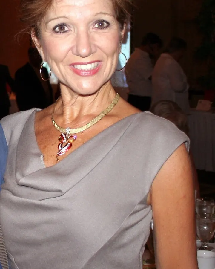 Femme aux cheveux châtains courts, souriante, portant une robe grise asymétrique et un collier orné d'un cœur multicolore.