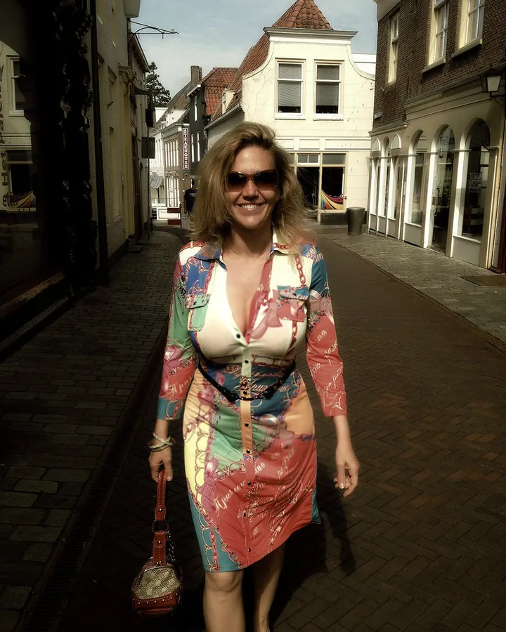 Femme aux cheveux blonds mi-longs, souriante, portant une robe colorée à motifs, marchant dans une rue pavée sous un soleil éclatant.