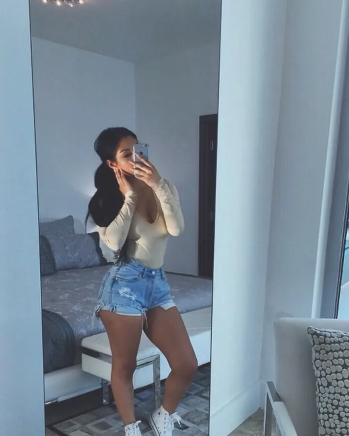 Femme en haut beige et shorts déchirés, prenant un selfie devant un miroir dans une chambre moderne