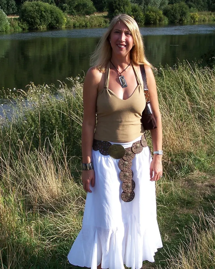 Femme en robe blanche et haut marron, debout près d'un lac, sourire, ceinture décorative