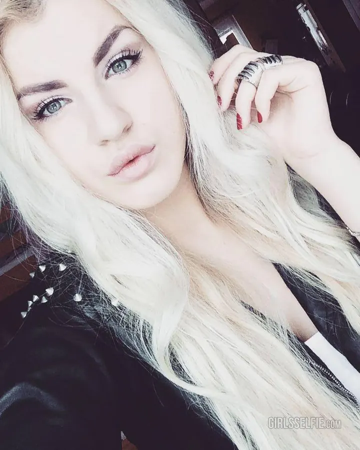 Jeune femme blonde aux yeux bleus, en tenue sombre, prenant un selfie avec une main dans les cheveux