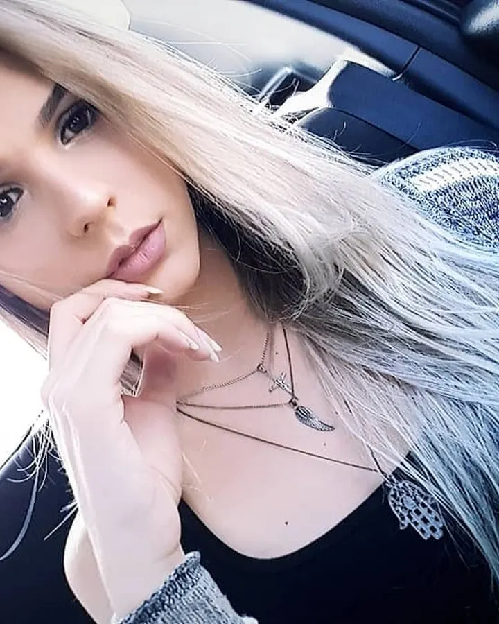 Jeune femme blonde, en pull gris et top noir, assise dans une voiture, regardant la caméra.