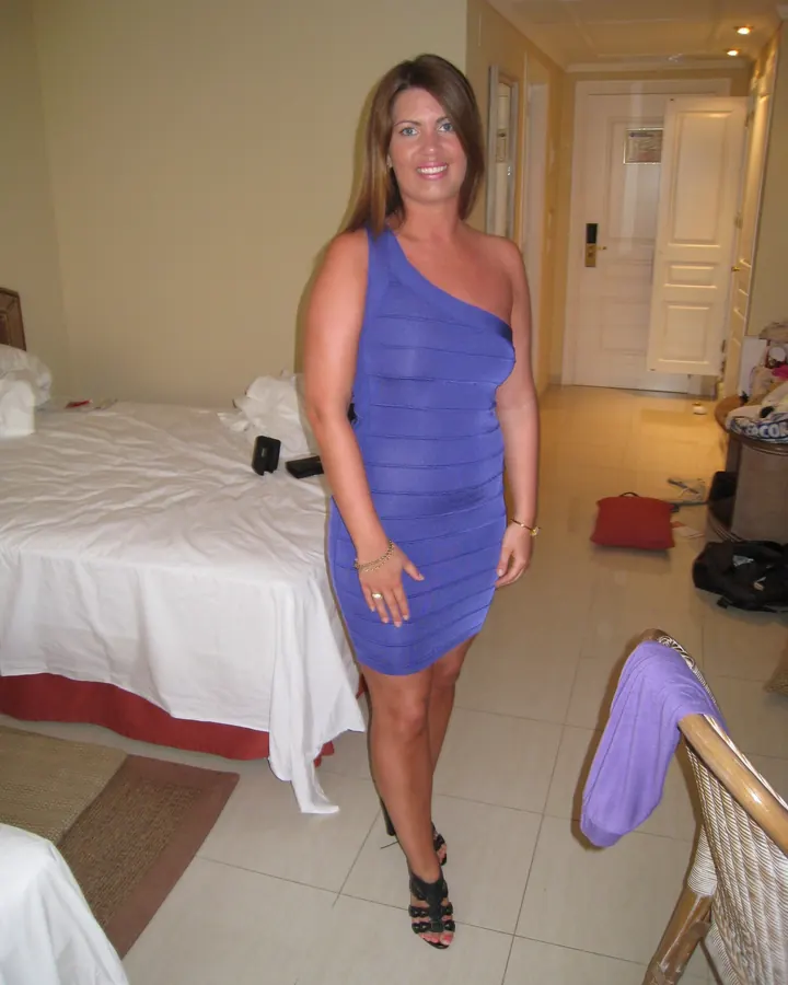 Femme blonde, en robe bleue asymétrique, posant dans une chambre d'hôtel.