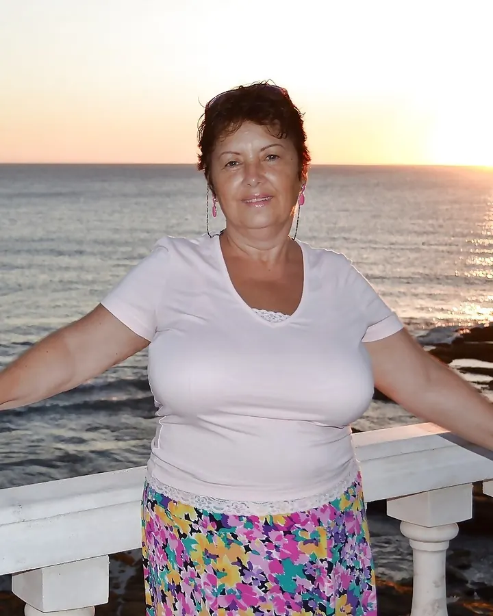Femme aux cheveux roux, souriante, en t-shirt blanc et jupe fleurie, devant la mer au coucher du soleil.