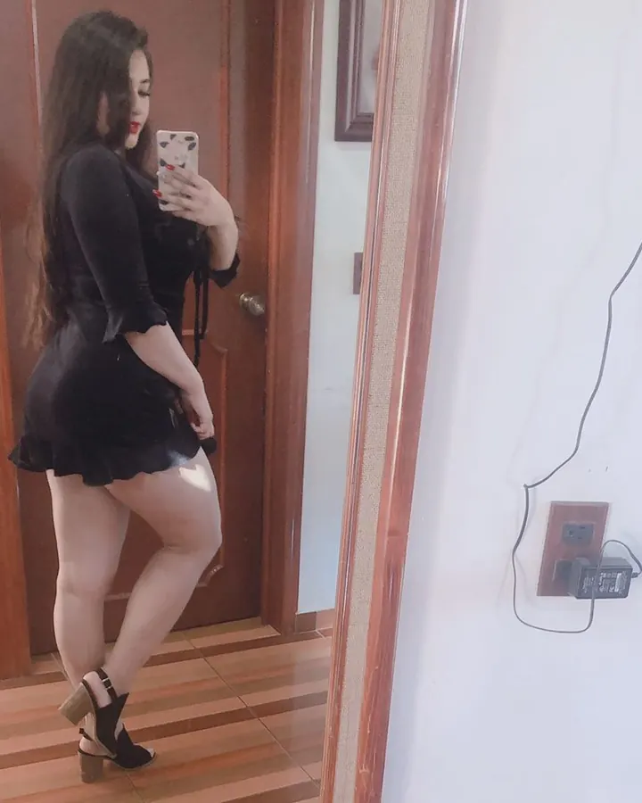 Femme aux cheveux bruns, en robe noire courte, prenant un selfie devant un miroir, dans une chambre.