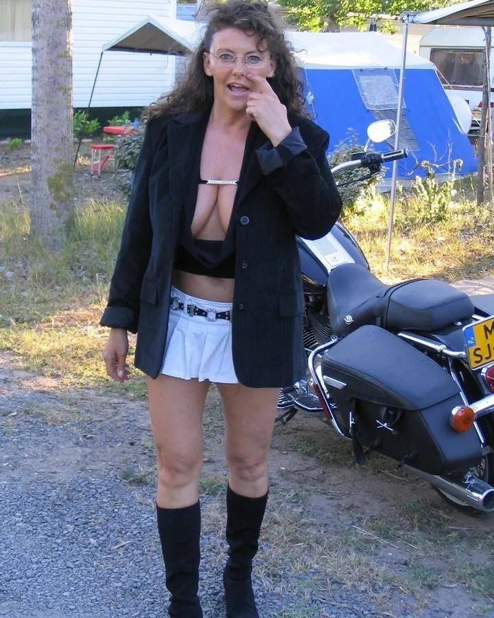 Femme aux cheveux bruns, en blazer noir et mini-jupe blanche, debout près d'une moto, avec des lunettes de vue.