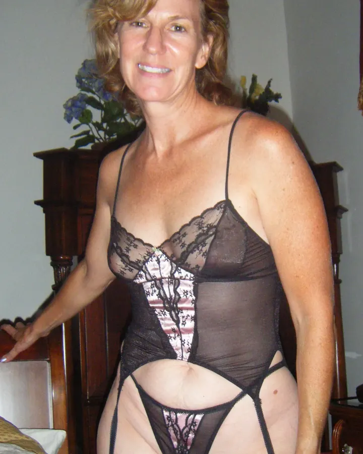 Femme aux cheveux blonds, en lingerie noire transparente, debout près d’un lit en bois, souriante.