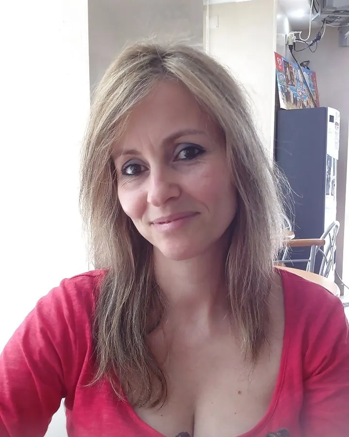 Femme aux cheveux gris, en chemisier rouge, souriant légèrement, dans un intérieur lumineux.