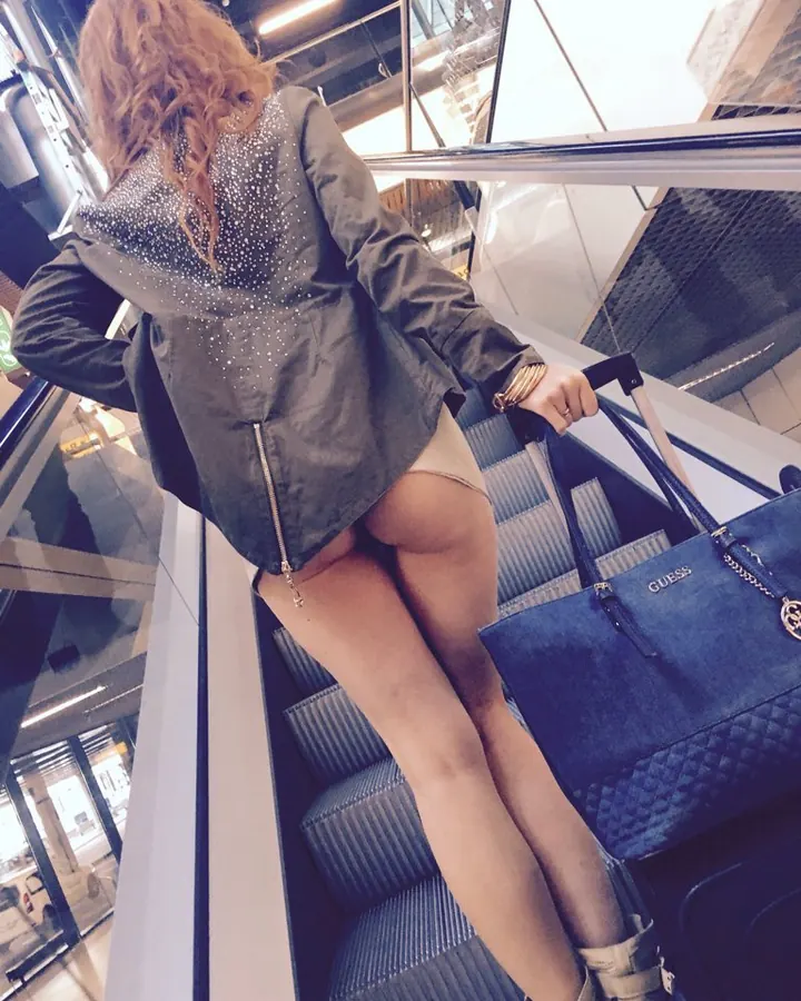 Femme aux cheveux châtains, en veste grise pailletée, montant un escalator avec une valise et un sac Guess.
