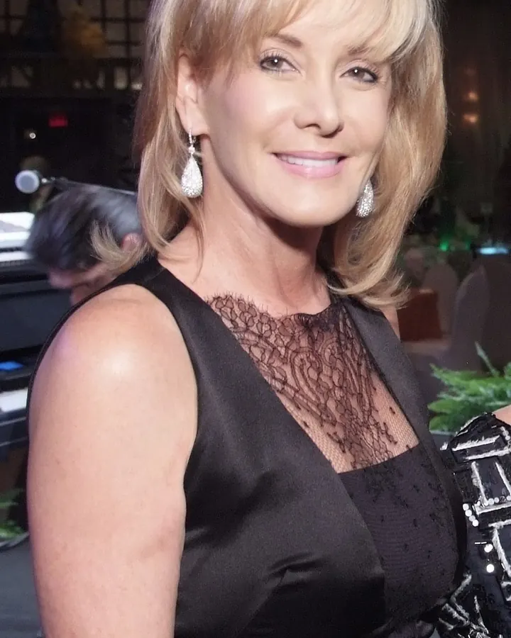 Femme blonde d'âge mûr, en robe noire avec dentelle, souriant légèrement à l'appareil.