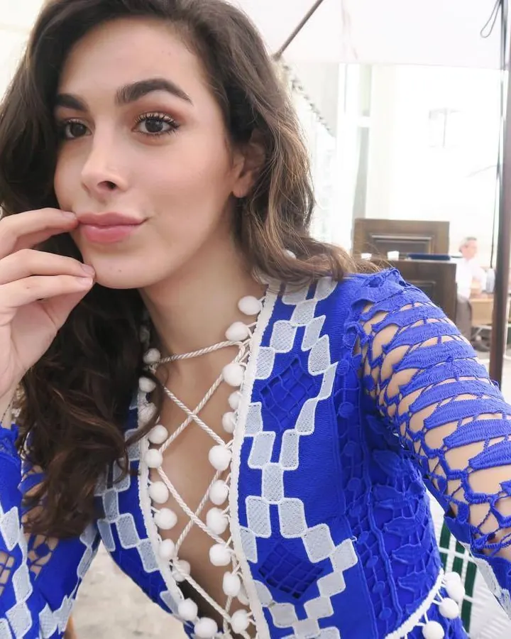 Jeune femme aux cheveux foncés, en robe bleue transparente, posant en extérieur, main sur la bouche.