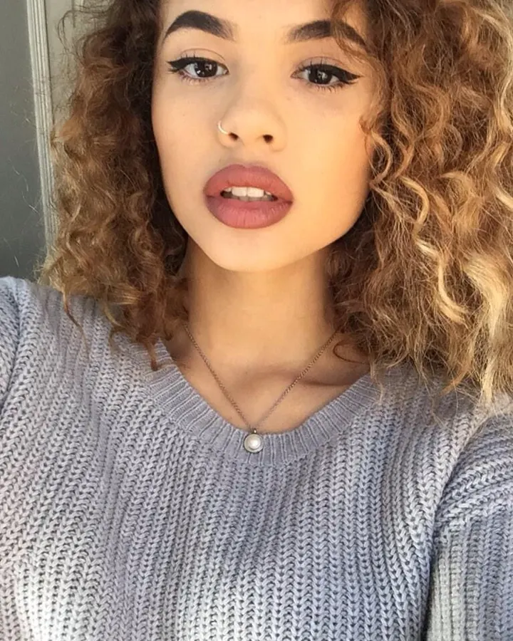 Femme aux cheveux blonds, portant un haut marron et des collants rouges, accroupie dans une cuisine.