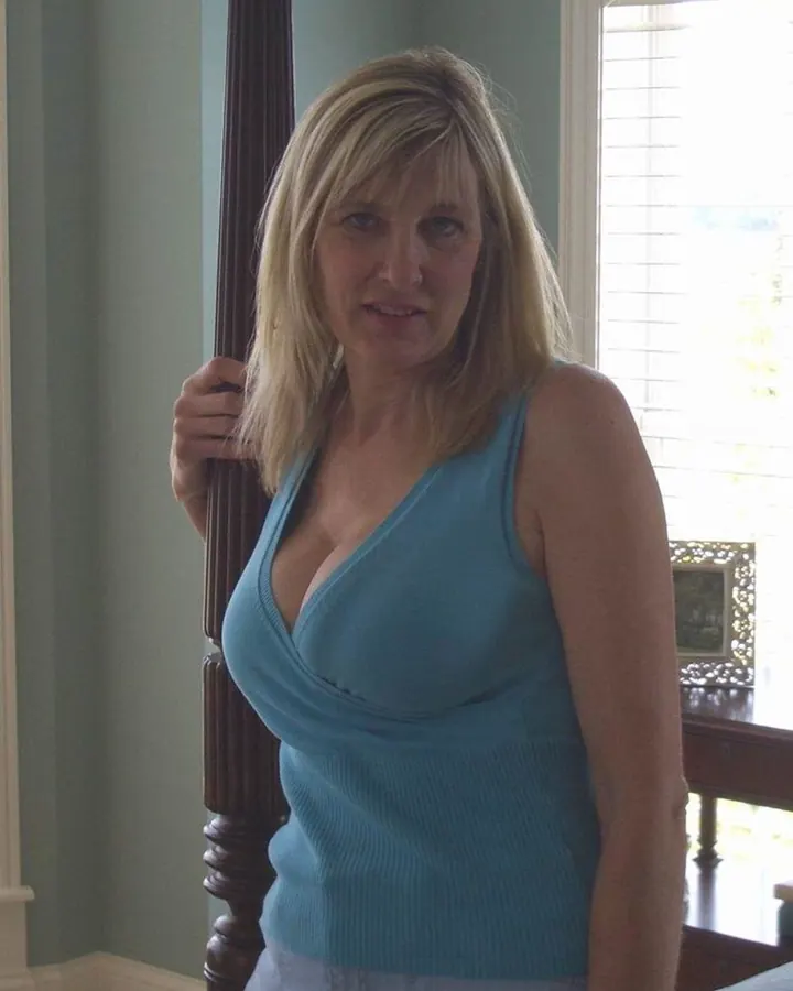 Femme aux cheveux blonds, portant un haut noir, assise sur un lit, tenant un téléphone.
