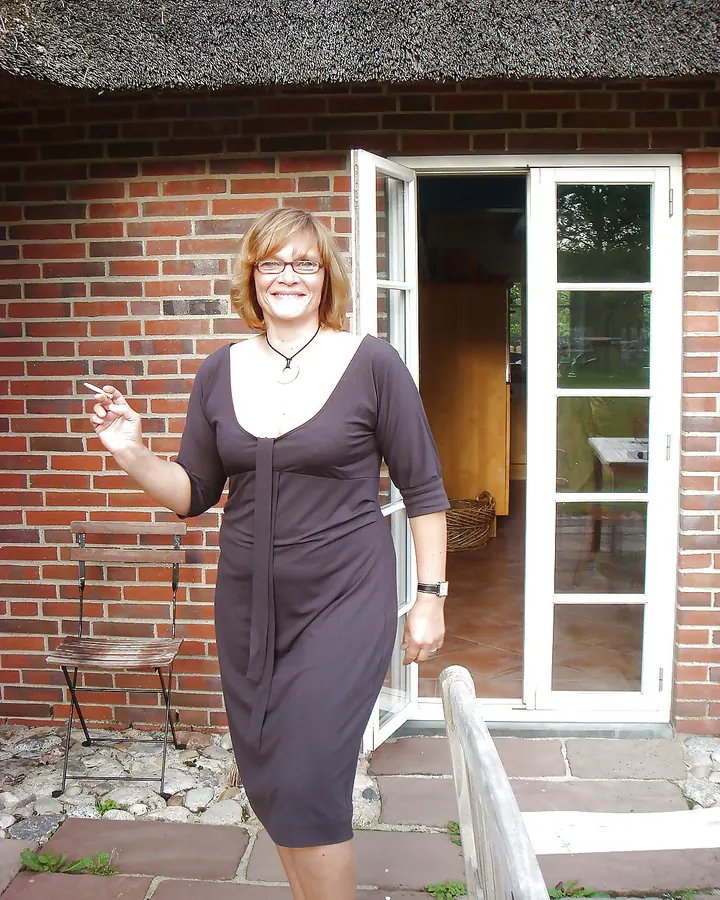 Femme blonde en robe marron, debout devant une maison en briques, tenant une cigarette