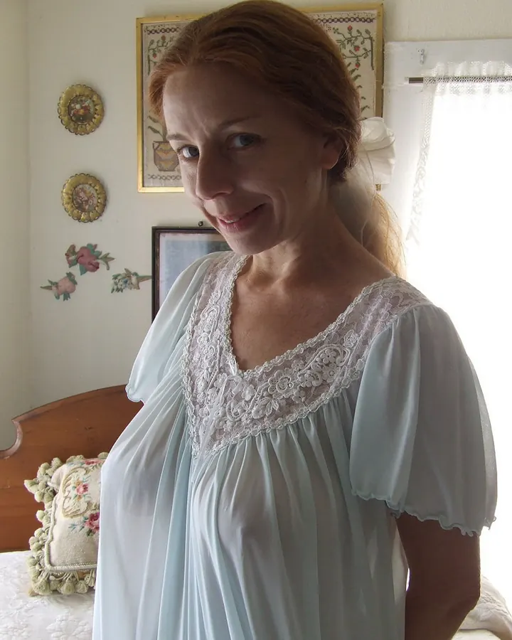 Femme blonde aux yeux gris, en robe bleu clair à dentelle, souriante, debout près d'un lit dans une chambre décorée de motifs floraux.