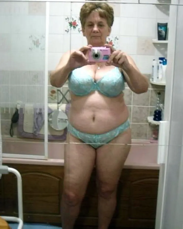 Femme âgée aux cheveux blonds courts, en sous-vêtements bleu clair, prenant un selfie dans une salle de bain devant un miroir.