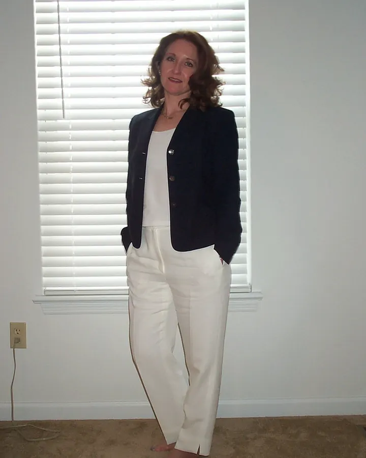Femme aux cheveux châtains, en chemise noire et pantalon blanc, debout devant une fenêtre aux volets fermés, pieds nus sur un tapis beige.